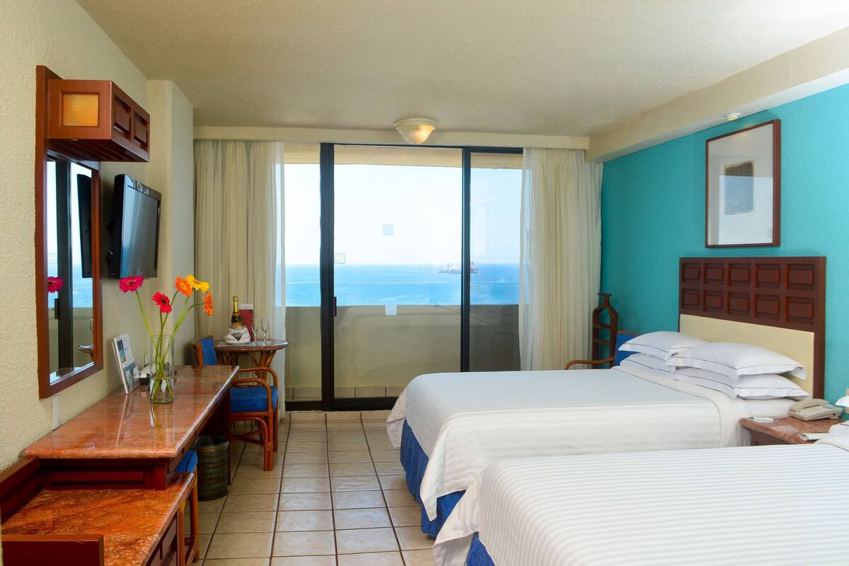 A hotel room with two beds, a desk with a chair, a TV, a window with a view of the sea, and a balcony.