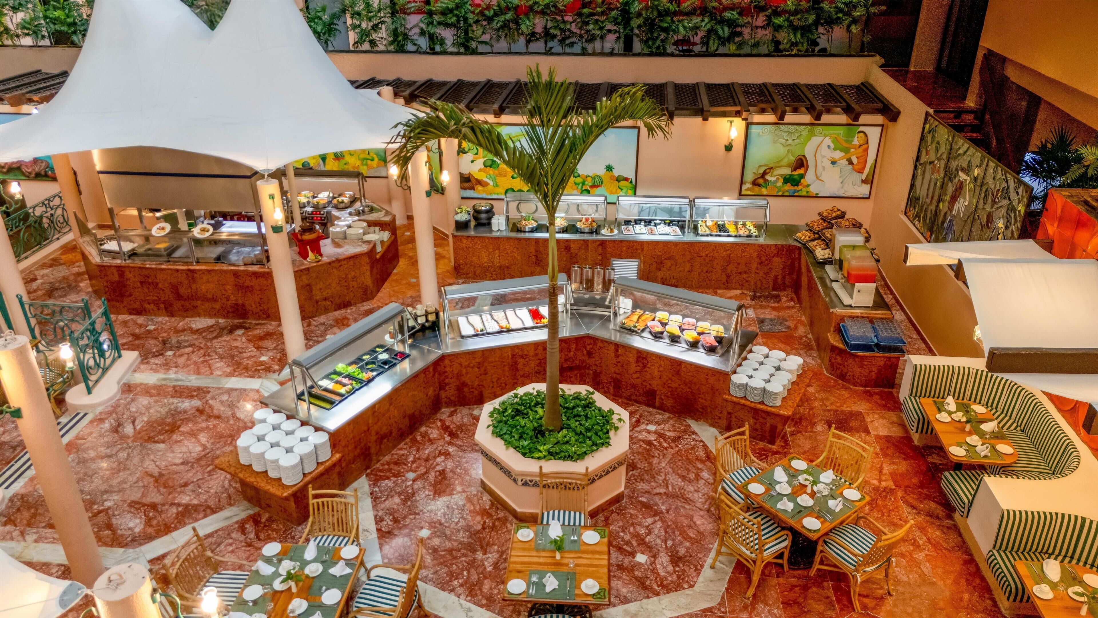 Un hall de hotel con una zona de estar central, varias mesas de buffet y una palmera decorativa.