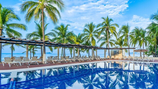 A pool with lounge chairs and palm trees, reflecting in the water. - Barceló Ixtapa All Inclusive (Ixtapa)