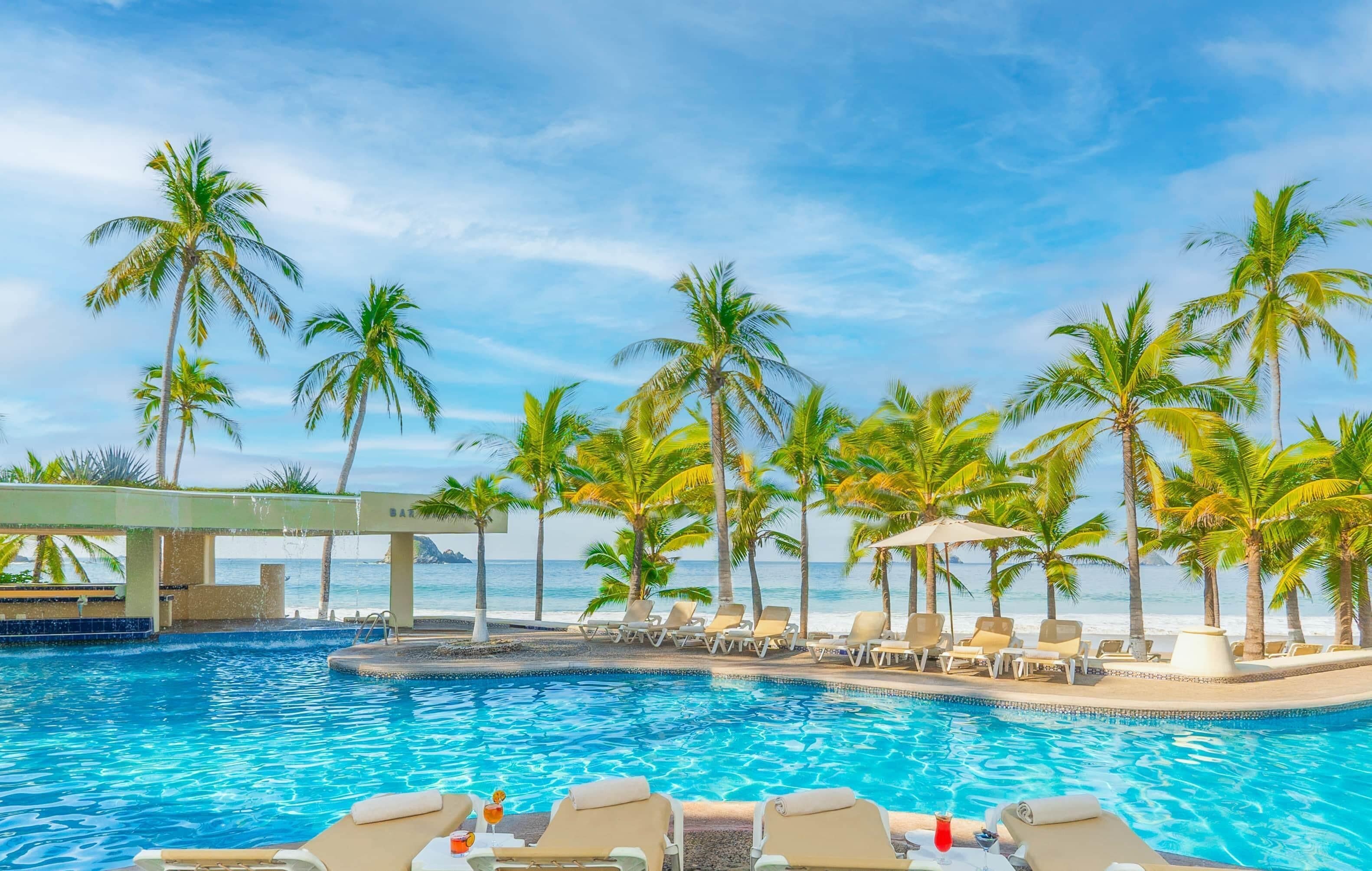 A pool area with lounge chairs and palm trees, overlooking a body of water.