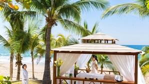 A beach cabana with a bed, a table with a vase of flowers, and a view of the ocean. - Barceló Ixtapa All Inclusive (Ixtapa)