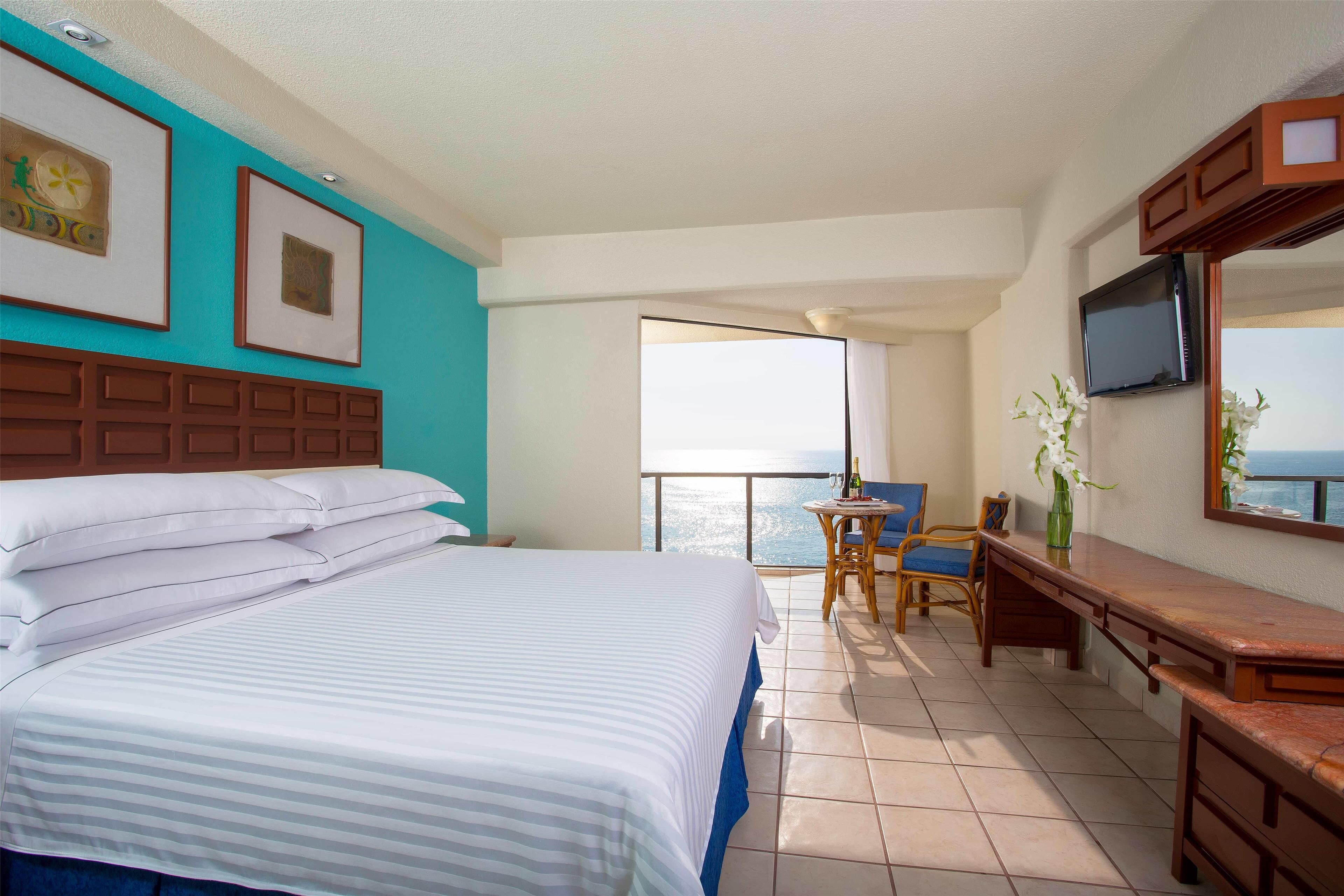 Una habitación de hotel con cama, escritorio, silla, televisor y balcón con vistas.