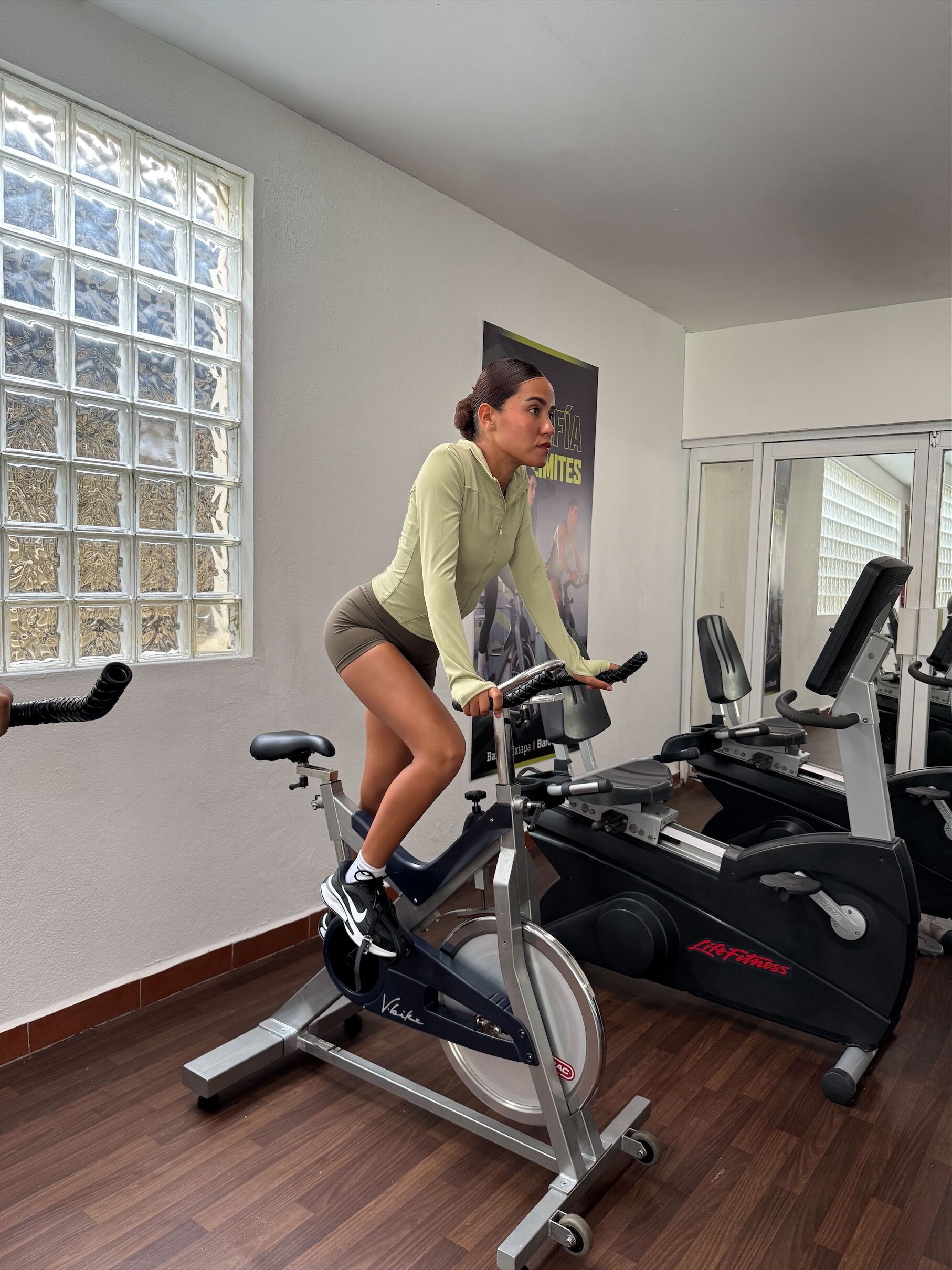 Una mujer usando una bicicleta estática en un gimnasio con una ventana, un cartel y equipo de ejercicio.