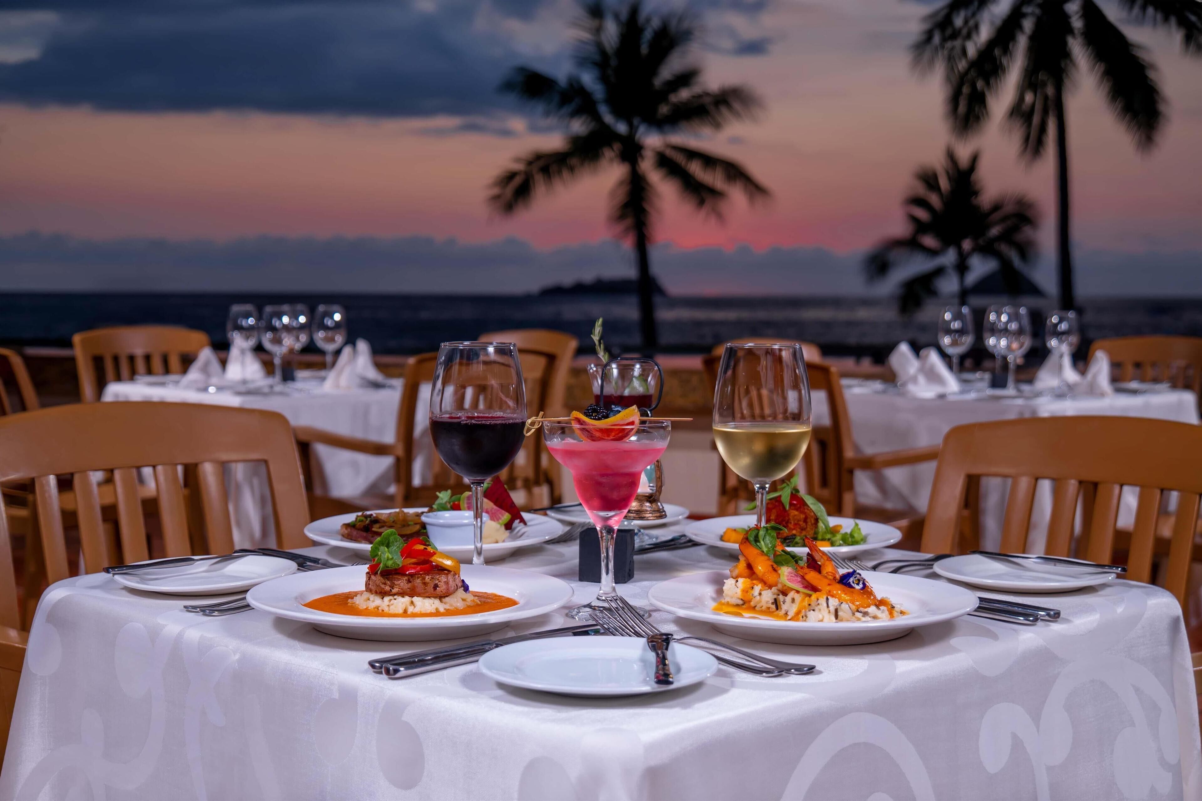 A table set for a meal with plates of food and glasses of wine, overlooking a beach with palm trees and a sunset.