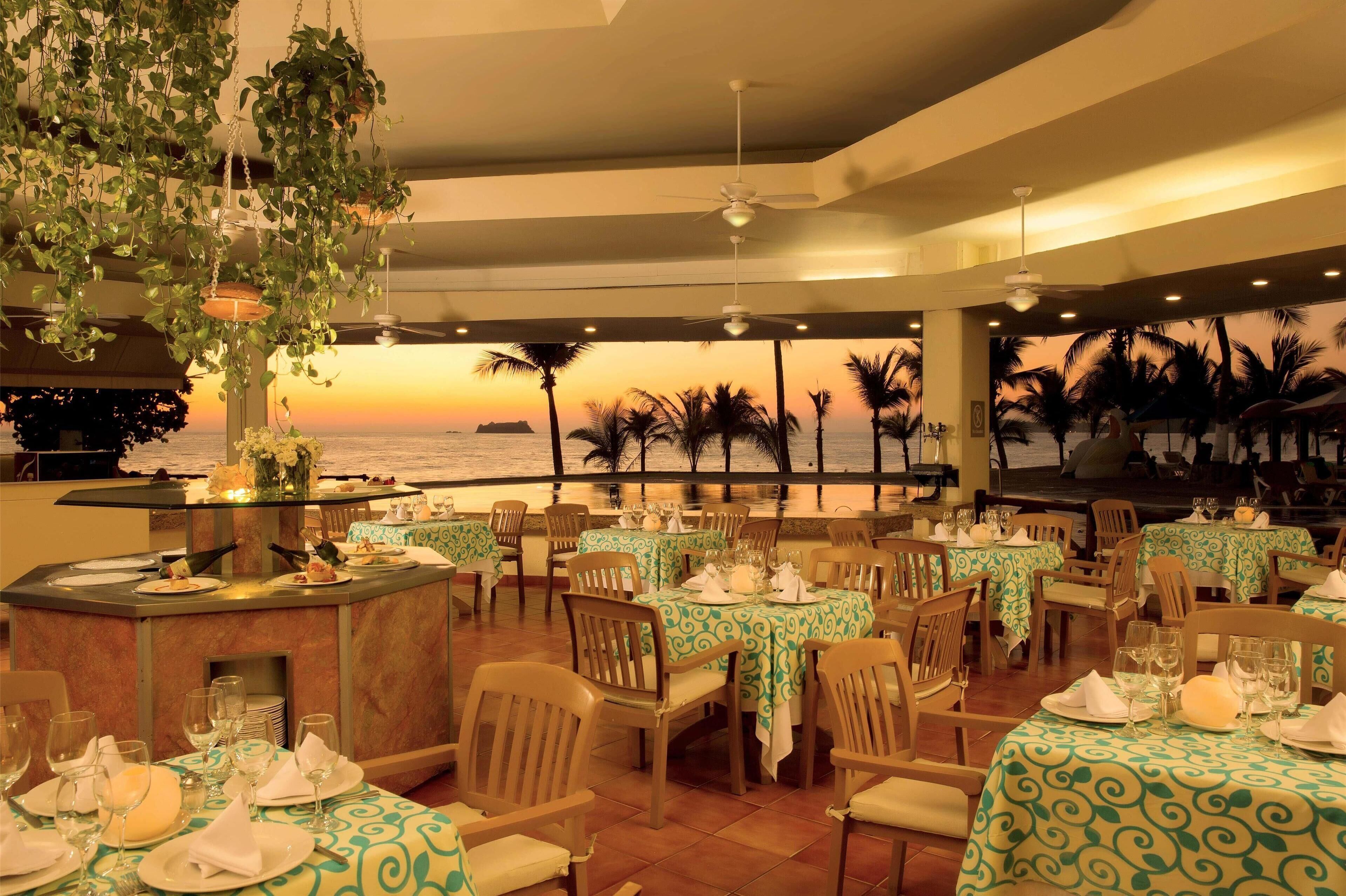 A restaurant with tables set for dining, a bar area, and a view of the sunset over the ocean.