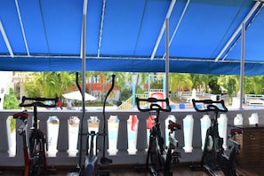 A covered outdoor area with exercise bikes and a view of a pool and buildings.