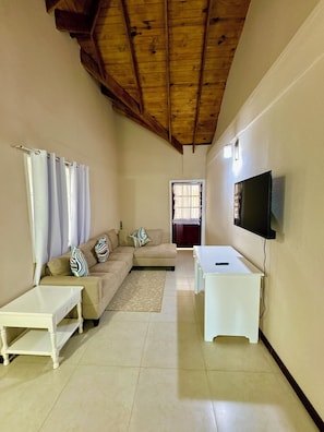 A living room with a wooden ceiling, a sofa with patterned cushions, a white coffee table, and a flat-screen TV mounted on the wall.