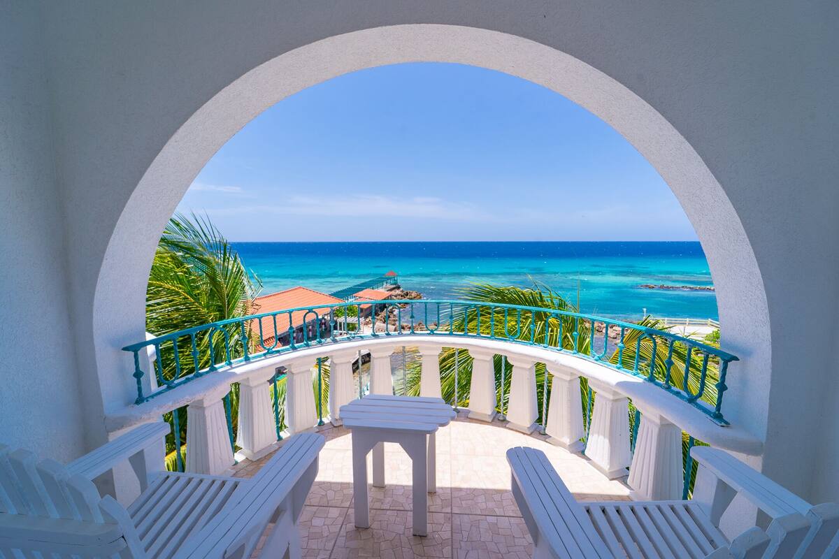 Un balcón con vista al océano, muebles blancos y barandilla turquesa.