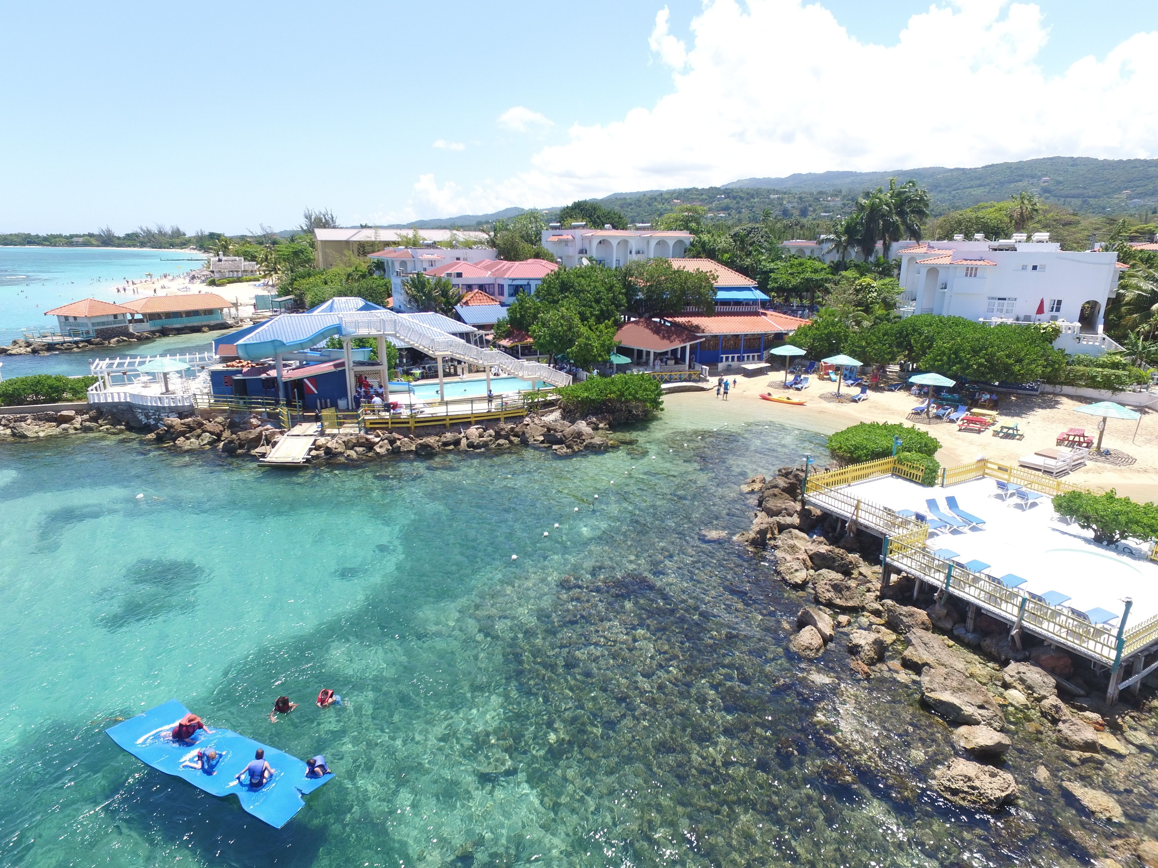 A coastal resort with clear waters, a beach, and several buildings with red roofs.