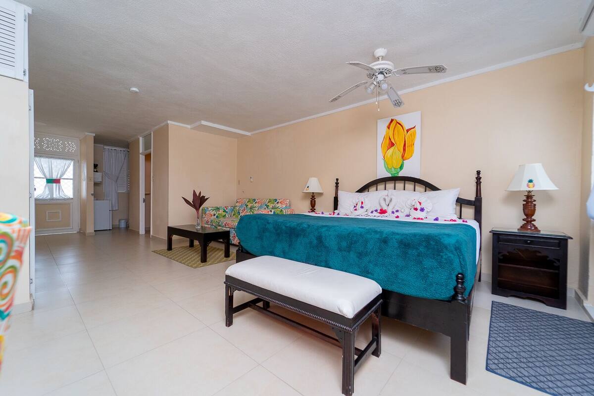 Un dormitorio con una cama, un banco, una mesita de noche, una lámpara, un ventilador de techo y un cuadro en la pared.
