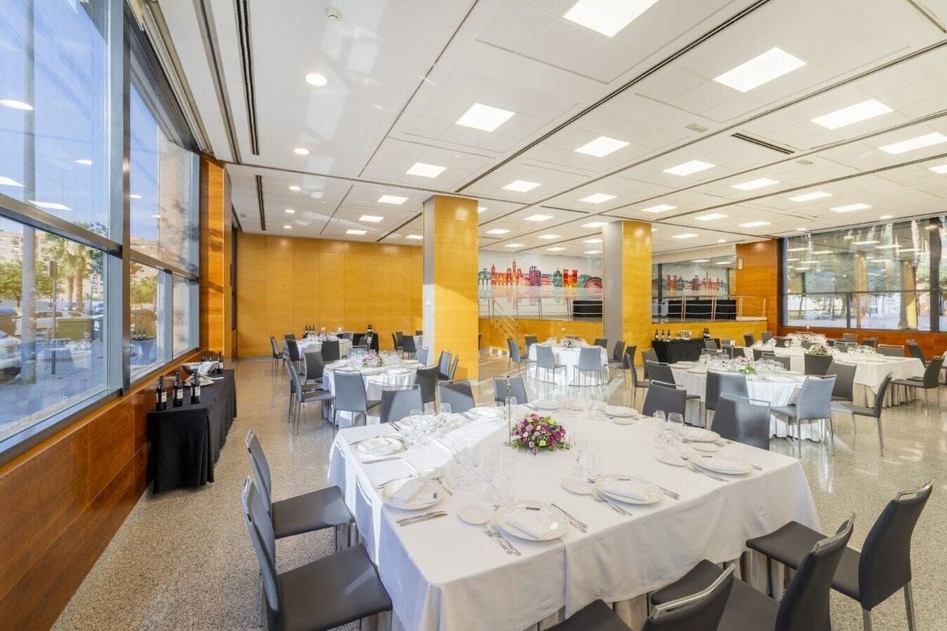 En rummelig festlokale med flere runde borde dækket til spisning, store vinduer med udsigt over byen og et moderne loftdesign.