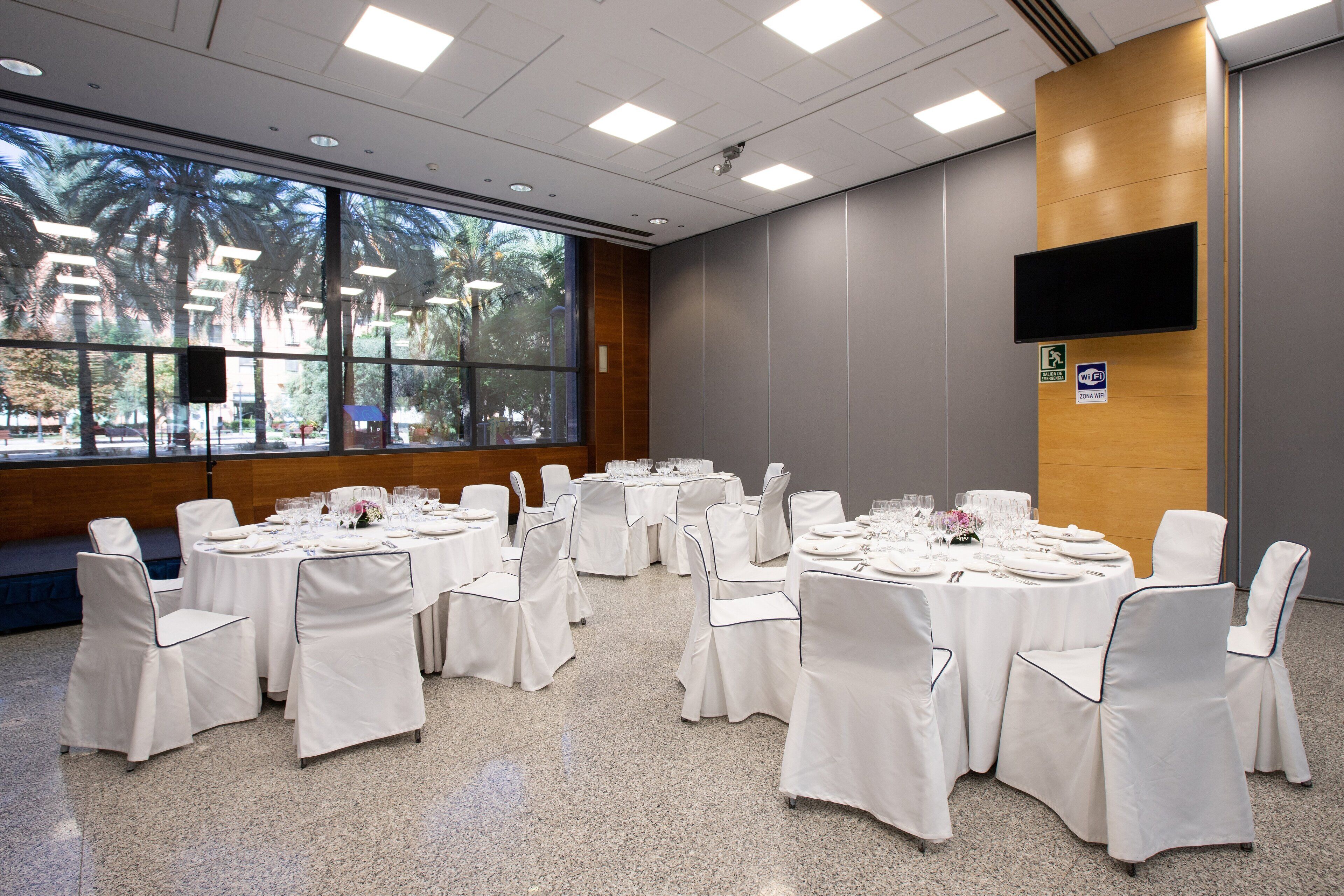 A conference room with round tables, white chairs, and a large window with a view of palm trees.