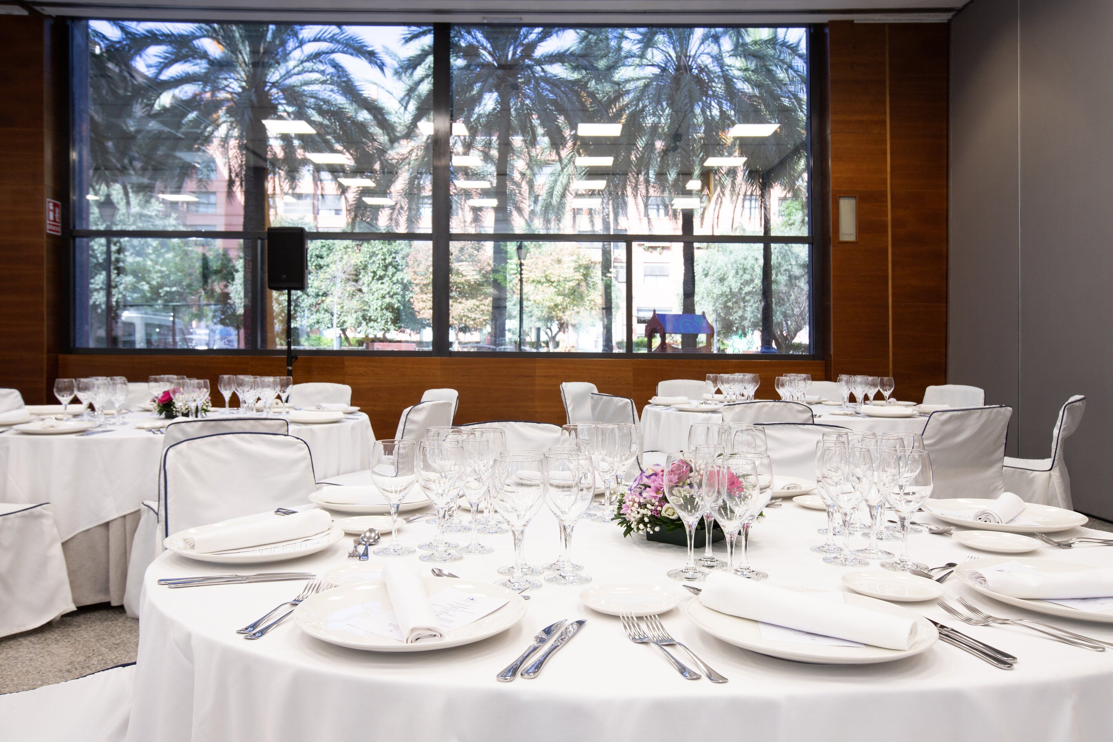 Un salón de conferencias con mesas redondas preparadas para un evento formal, grandes ventanales con vista a palmeras y un altavoz montado en la pared.