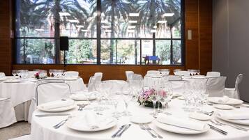 Un salón de conferencias con mesas redondas preparadas para un evento formal, grandes ventanales con vista a palmeras y un altavoz montado en la pared.