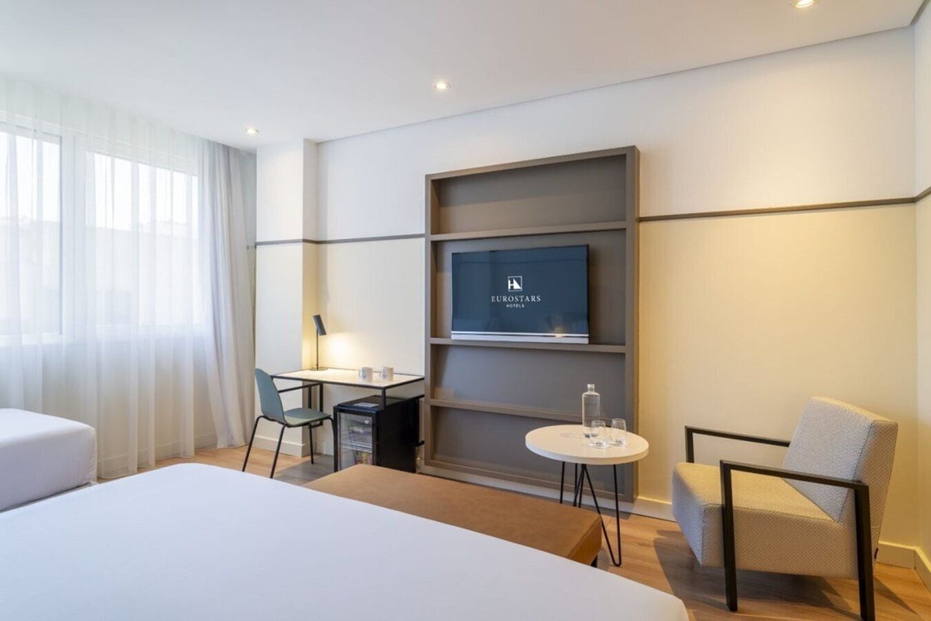 A modern hotel room with a large bed, a desk with a chair, a small table with a chair, a TV on the wall, and a window with sheer curtains.