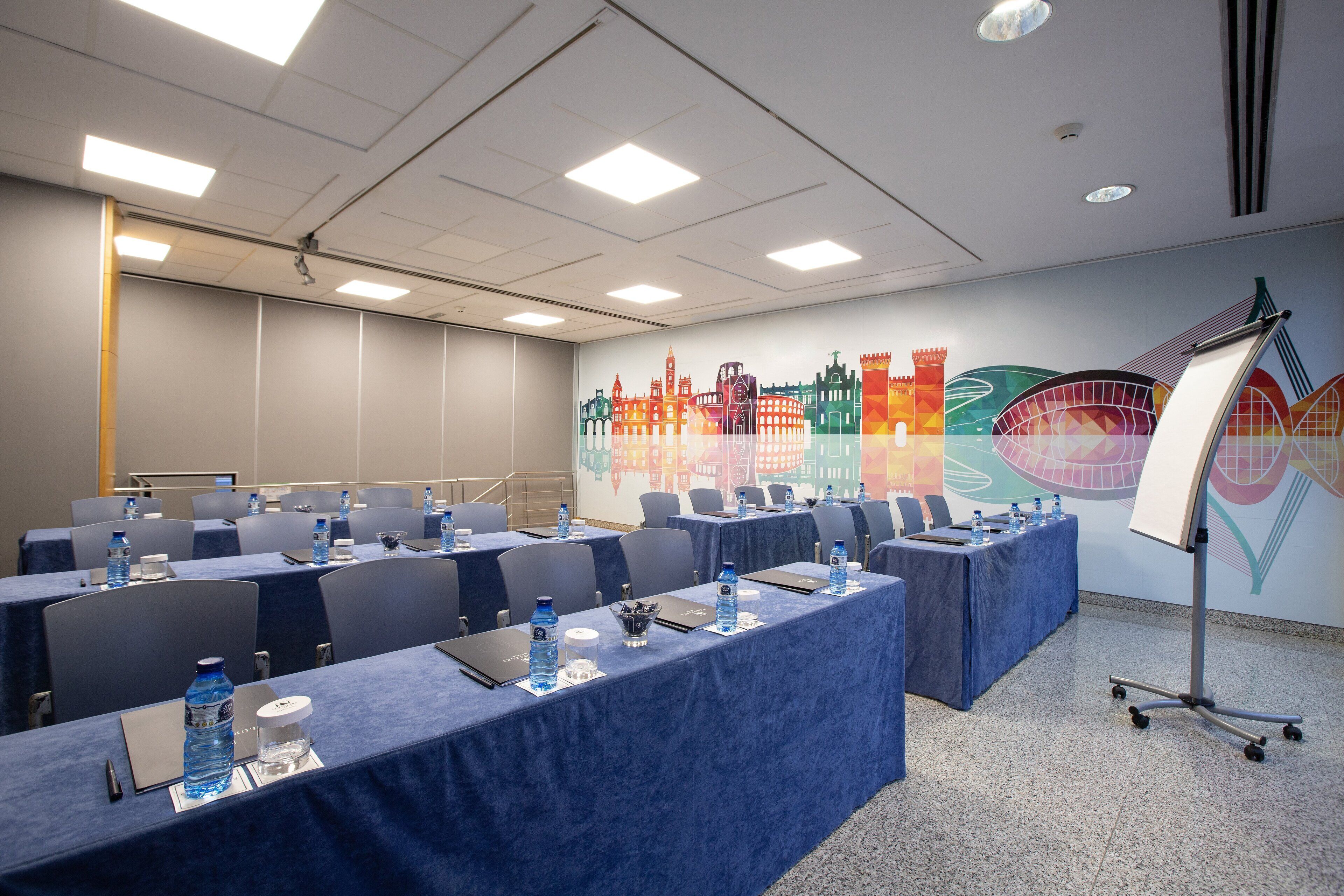 Une salle de conférence avec des tables disposées pour une réunion, équipée d’ordinateurs portables, de bouteilles d’eau et de blocs-notes.