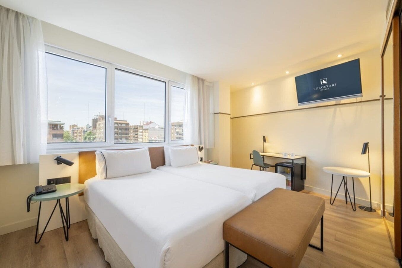 A hotel room with a large bed, a desk, a chair, and a view of the city through large windows.
