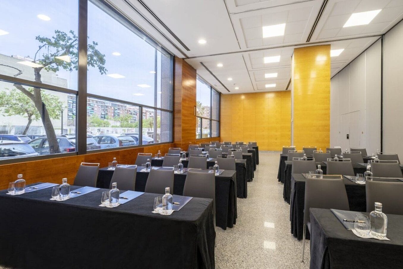 Un salón de conferencias con filas de mesas y sillas, ventanas amplias que dan vista a coches estacionados y edificios afuera.