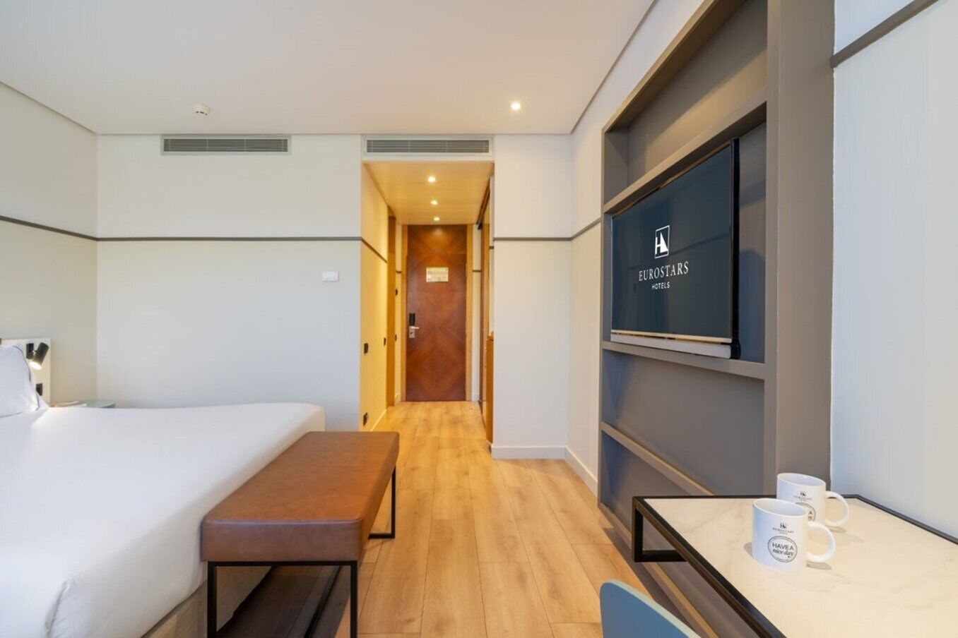 A hotel room with a bed, a bench, a desk, and a TV on the wall.