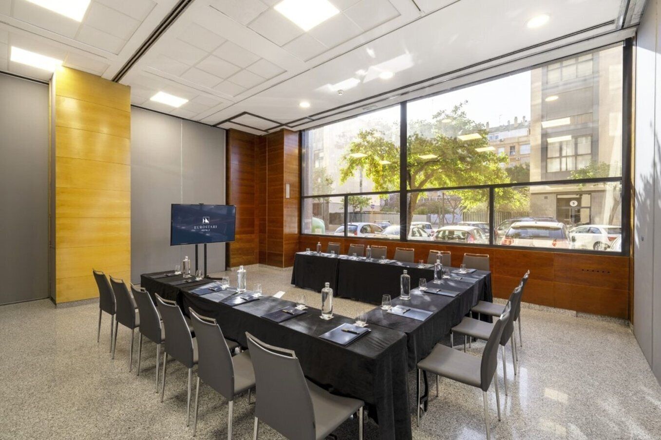 A conference room with a long table, chairs, a screen, and a view of parked cars and buildings through large windows.