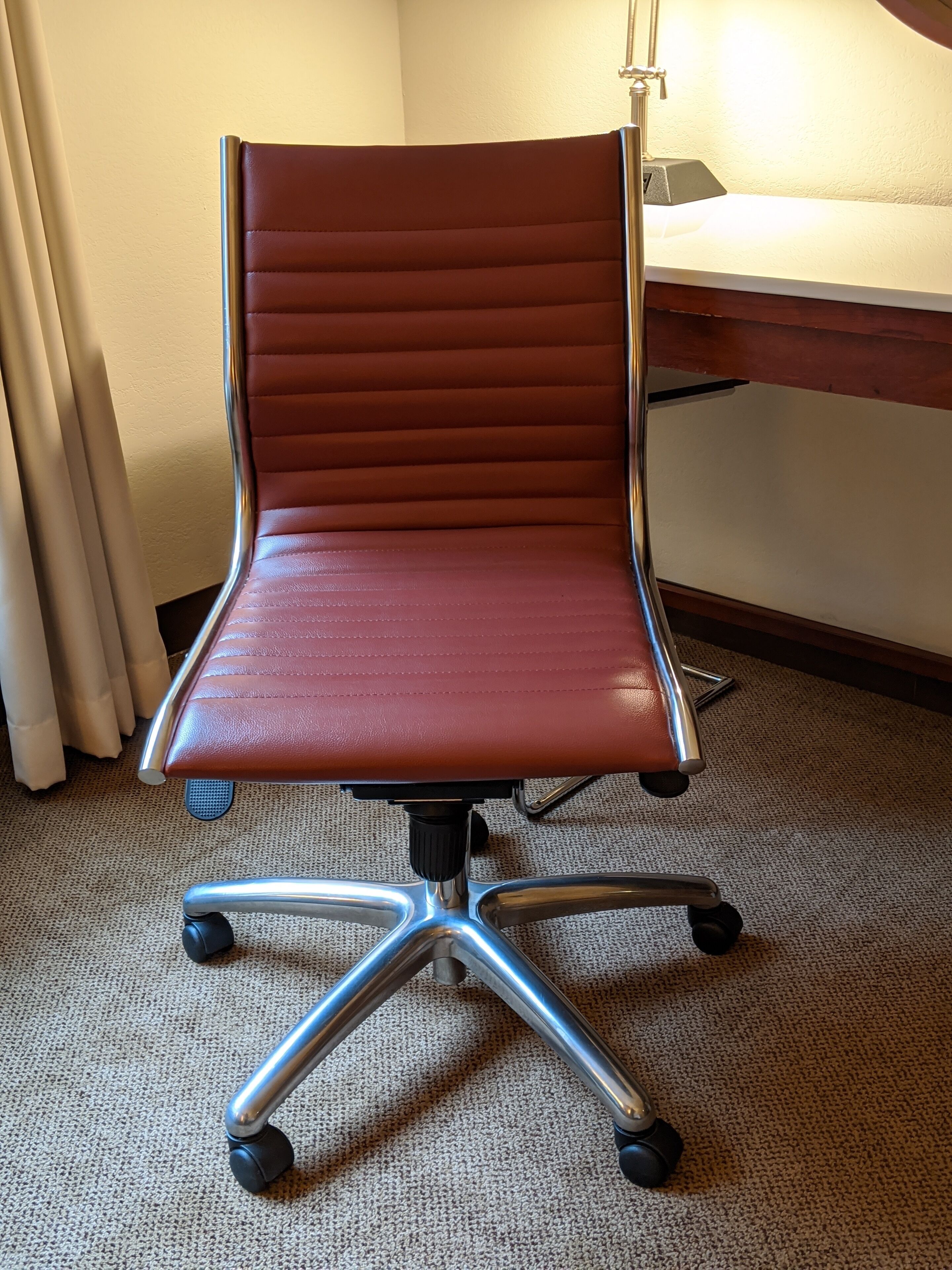 Ein roter Bürostuhl mit hoher Rückenlehne und einer Fünf-Speichen-Fußkreuzbasis steht auf einem Teppichboden in einem Raum mit einem Holztisch und einer Lampe.