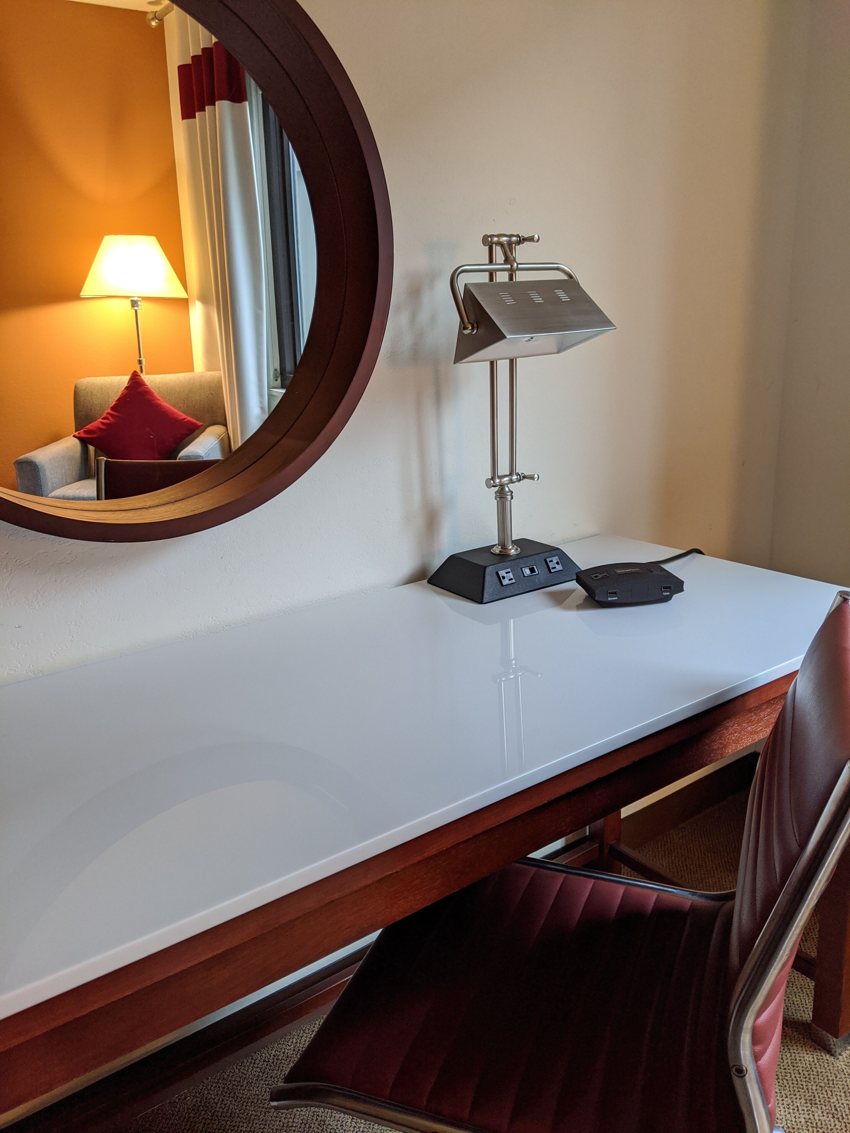 A hotel room with a round mirror, a desk with a lamp, and a chair.