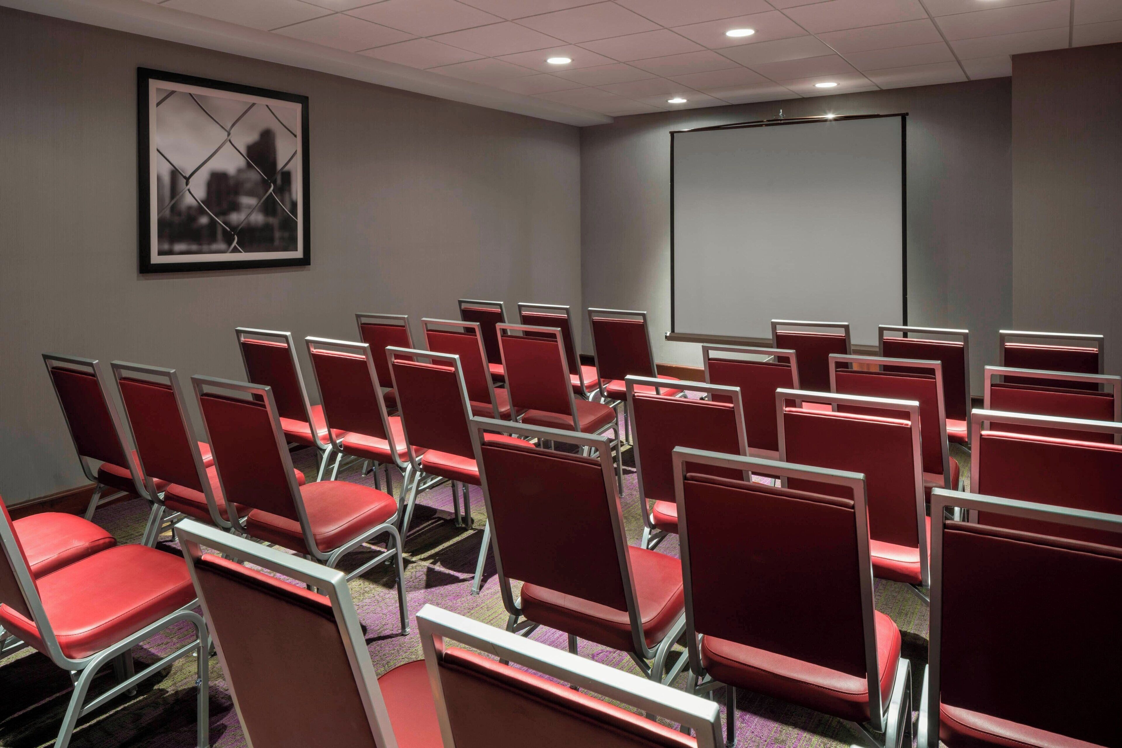 A conference room with rows of red chairs, a projector screen, and a framed picture on the wall.