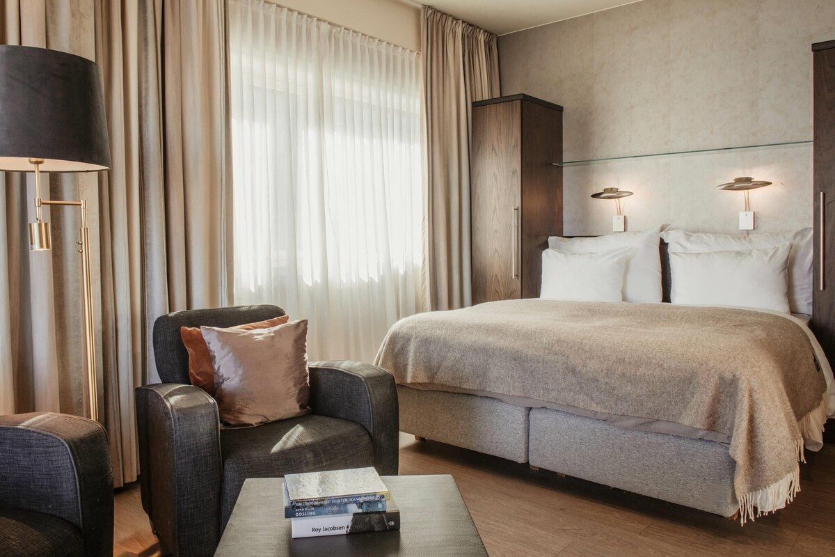 A hotel room with a bed, a sofa, a coffee table with books, a lamp, and curtains.