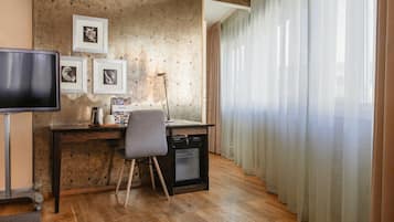A room with a wooden desk, a chair, a television, a refrigerator, and a window with sheer curtains.