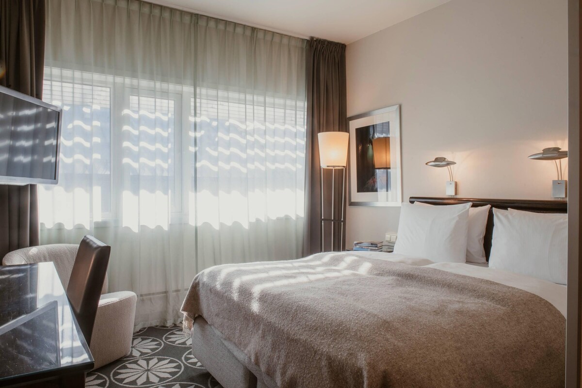 Habitación de hotel con cama, escritorio, silla, televisor, lámpara y ventana con cortinas.