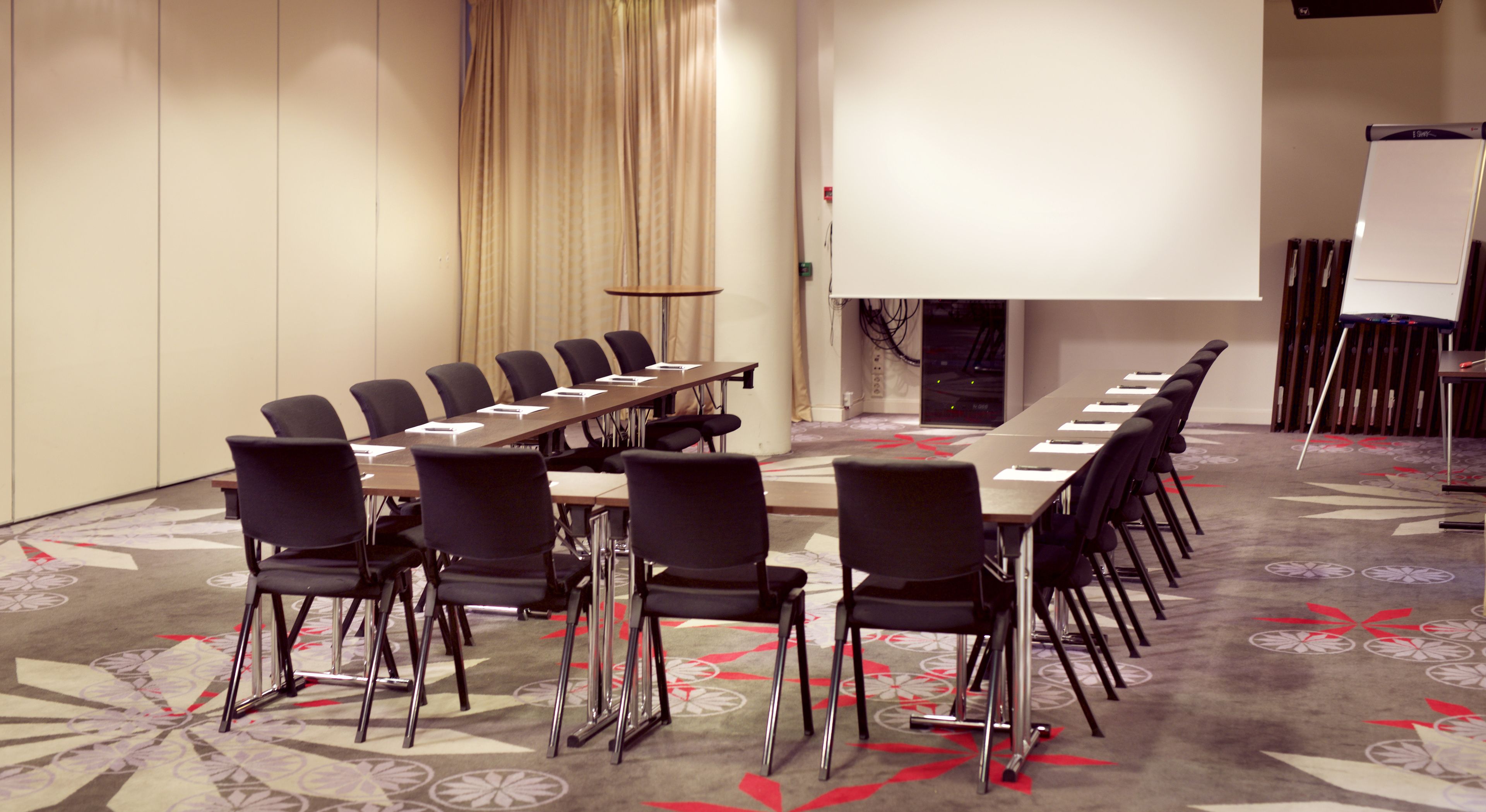 A conference room with a long table, chairs, a projector screen, and a carpeted floor.