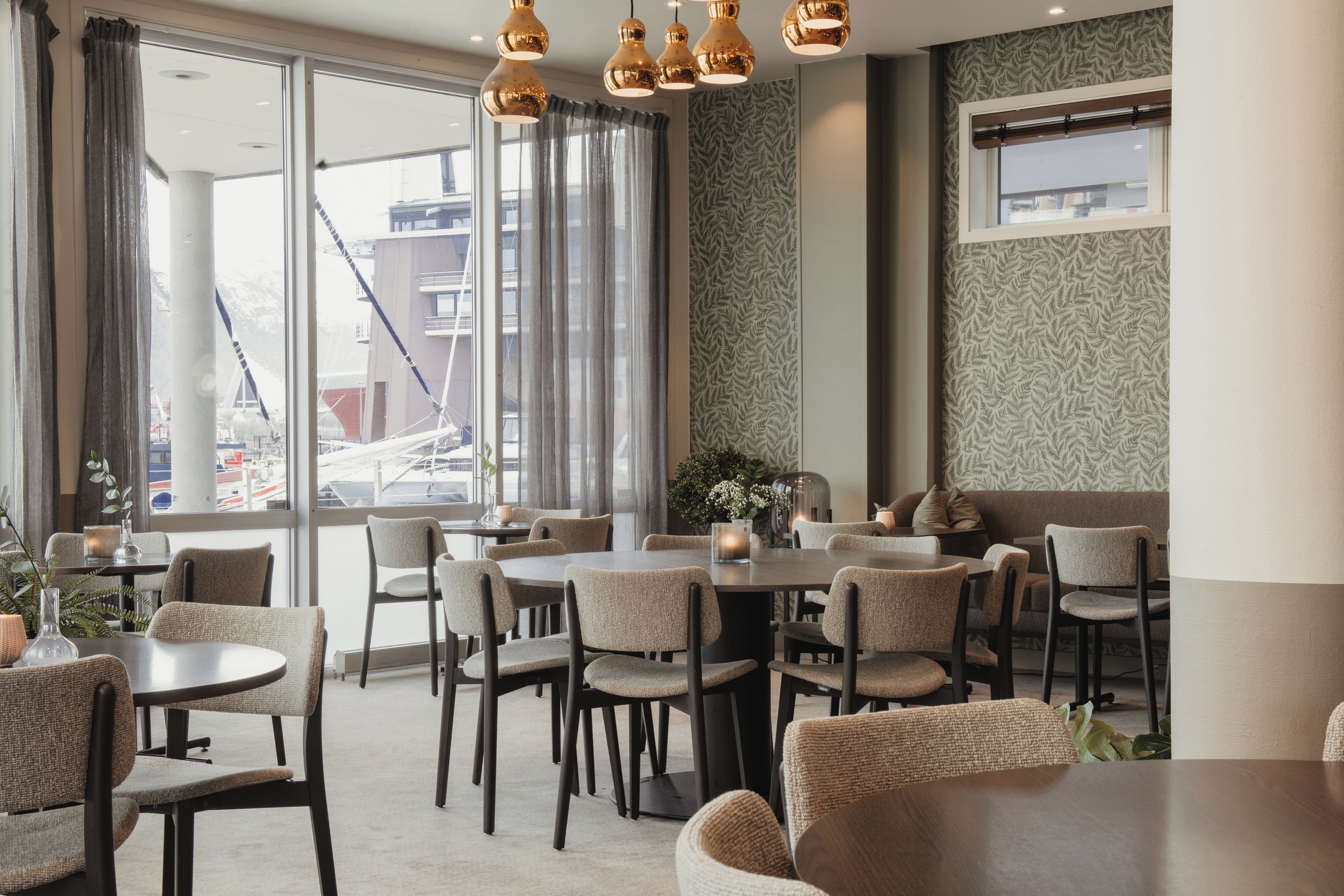 Un interior de restaurante con mesas y sillas redondas, ventanas grandes con cortinas y una pared con diseño.