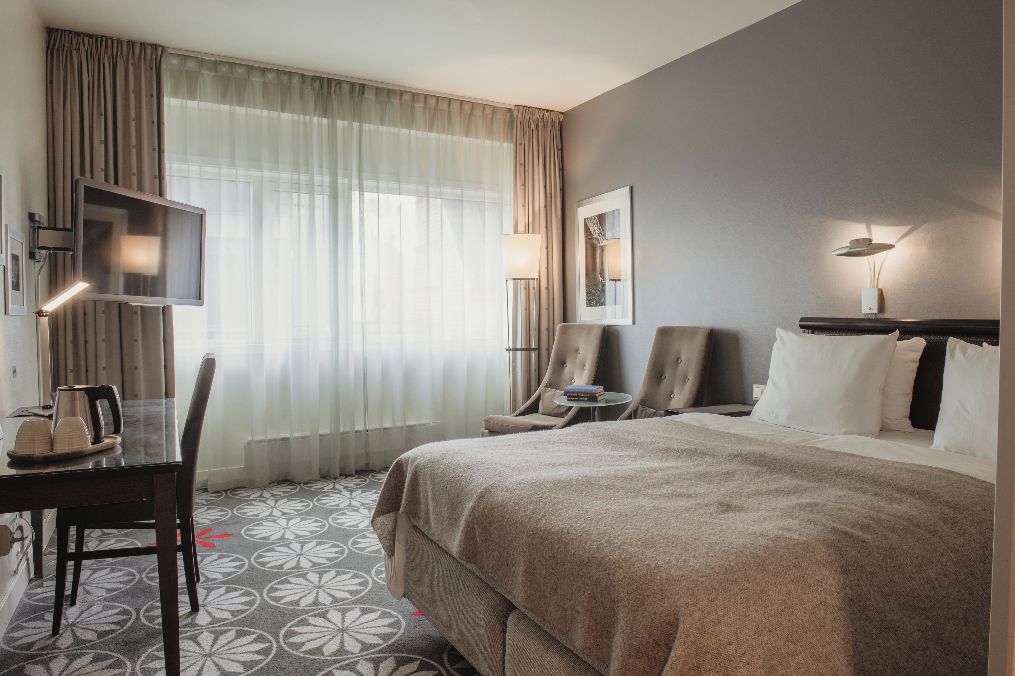 A hotel room with a bed, two chairs, a desk with a kettle, a flat-screen TV, and a large window with curtains.