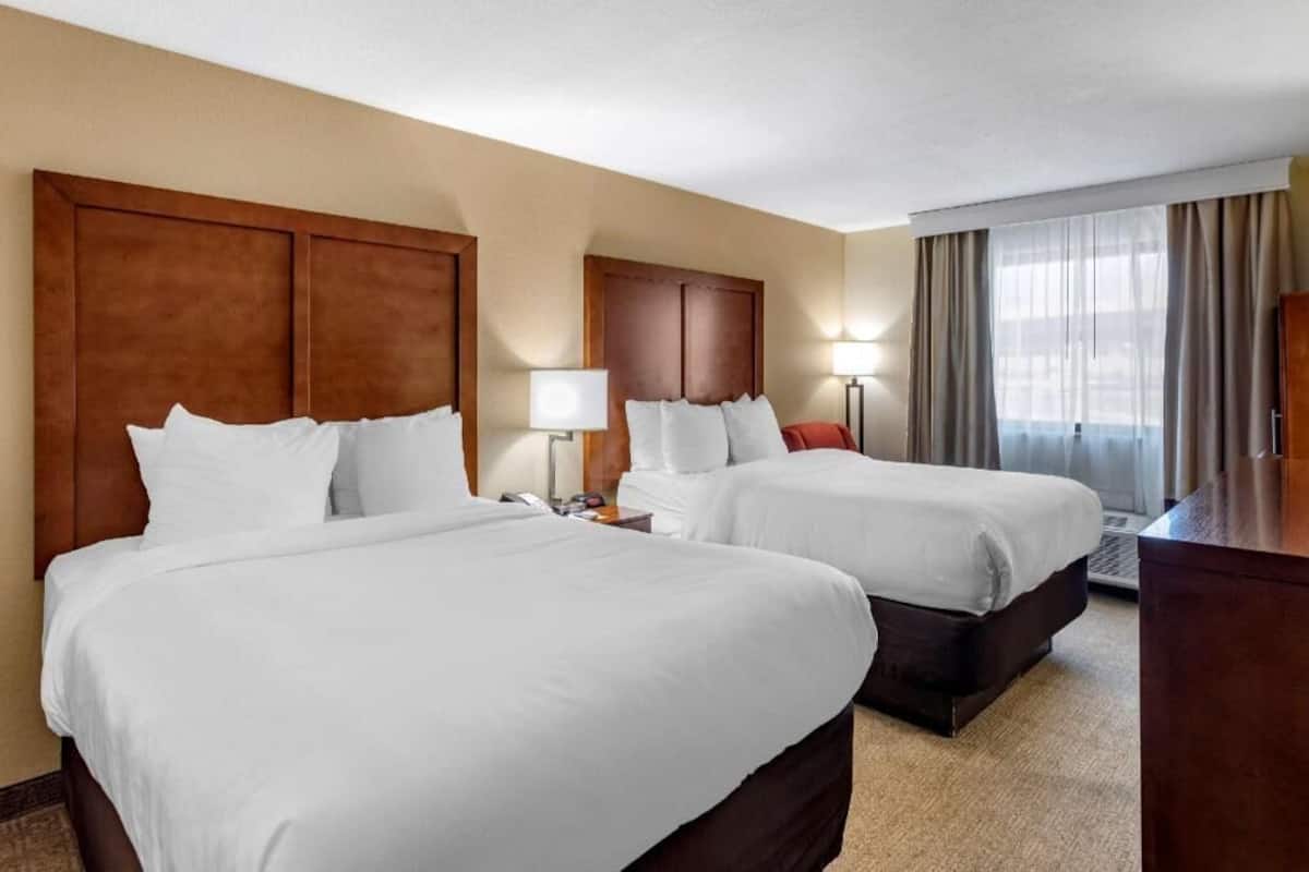 A hotel room with two beds, a desk, a lamp, and a view of the outdoors.