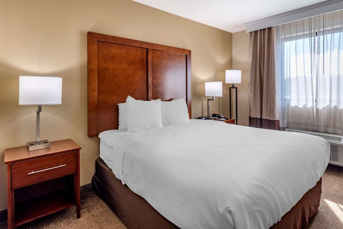 A hotel room with a large bed, wooden headboard, two bedside tables with lamps, and a window with curtains.