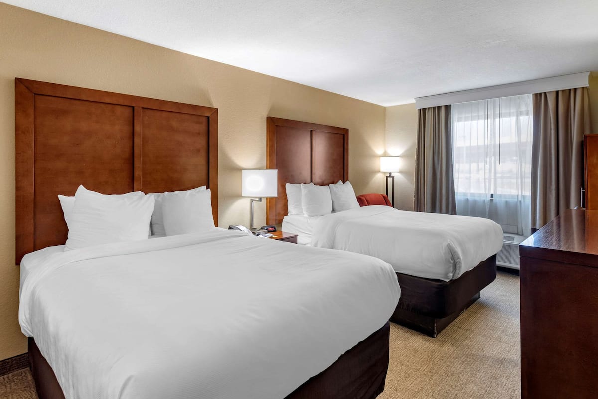 A hotel room with two beds, a wooden headboard, a nightstand with a lamp, a desk, and a window with curtains.