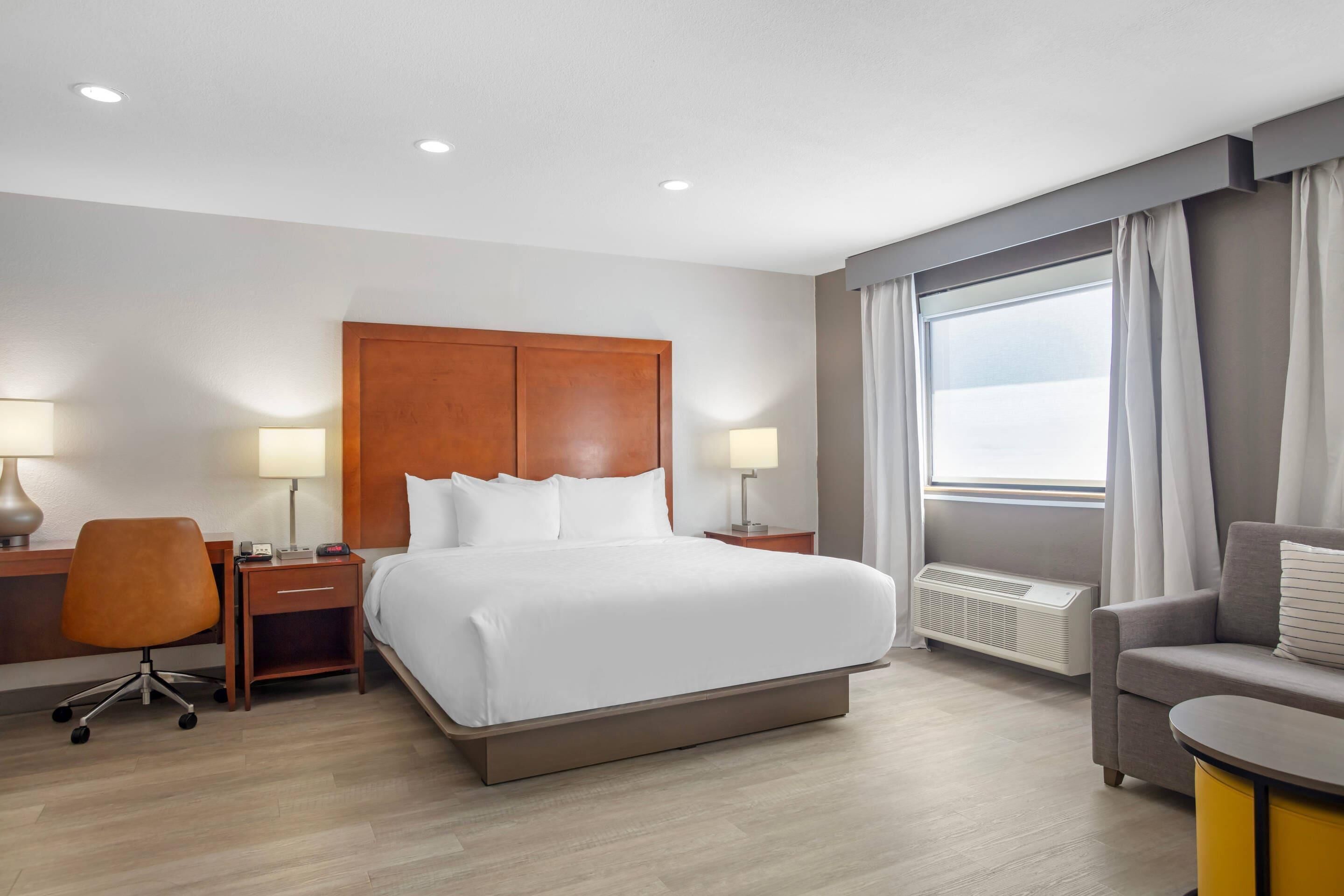 A hotel room with a large bed, a desk with a chair, a bedside table, a radiator, and a window with curtains.