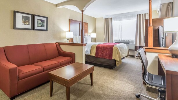 A hotel room with a red sofa, a wooden coffee table, a bed with a red blanket, a desk with a lamp, and two framed pictures on the wall. - Comfort Inn Nashville West (Nashville)