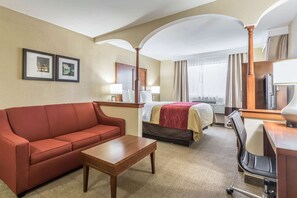 A hotel room with a red sofa, a wooden coffee table, a bed with a red blanket, a desk with a lamp, and two framed pictures on the wall.