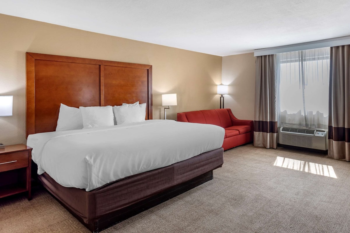 A hotel room with a large bed, a red armchair, a nightstand, a lamp, and a window with curtains.