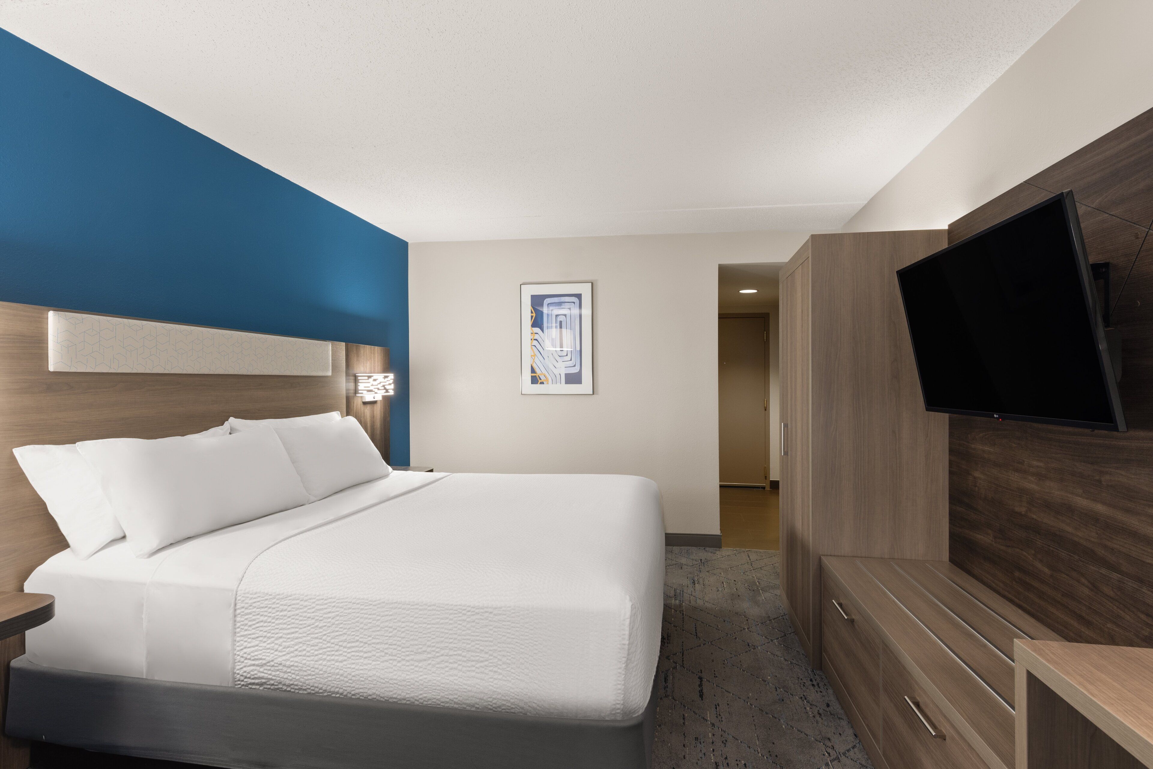 A modern hotel room with a large bed, a flat-screen TV mounted on the wall, and a wooden wardrobe.