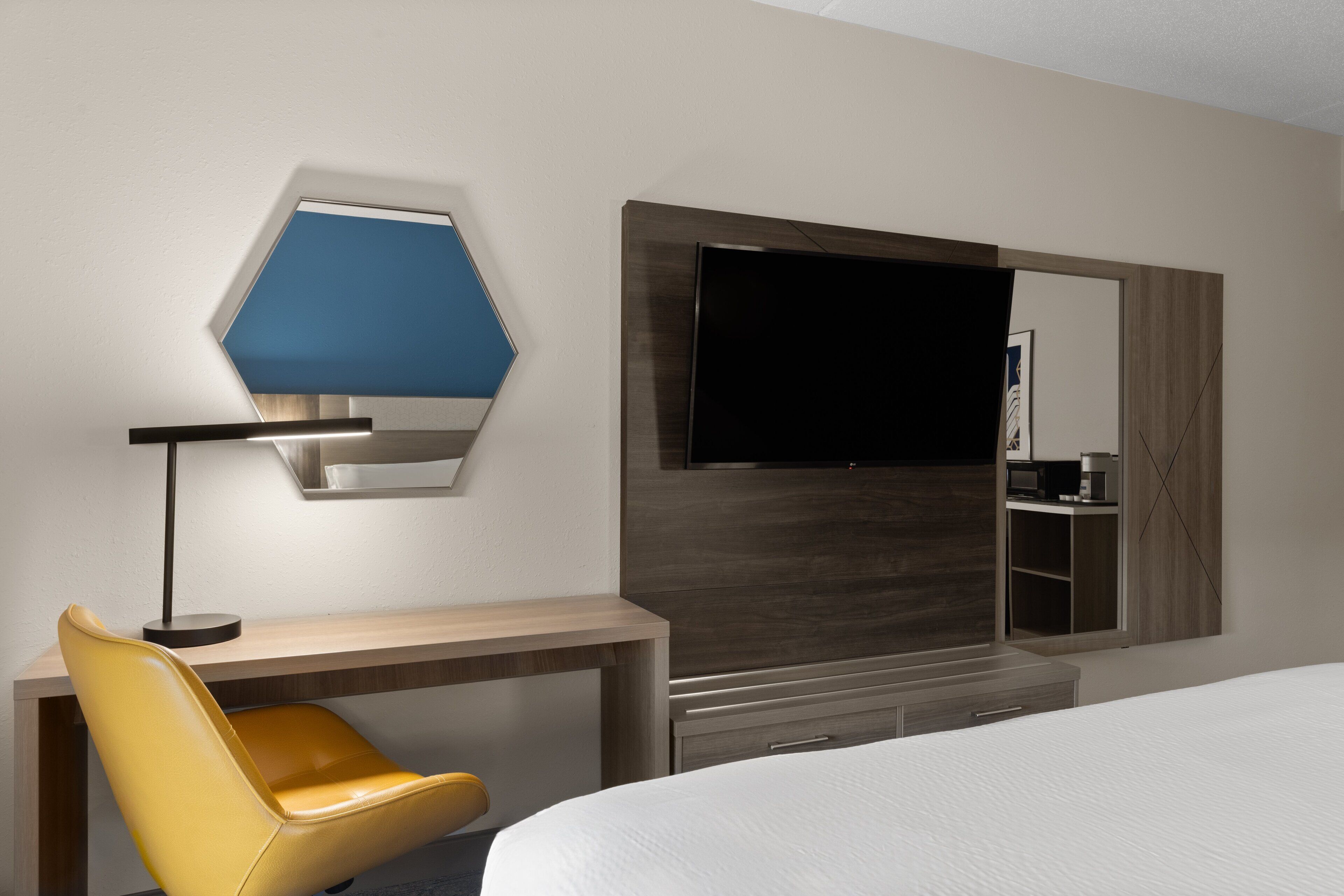 A hotel room with a bed, a desk with a chair, a flat-screen TV, and a large mirror.