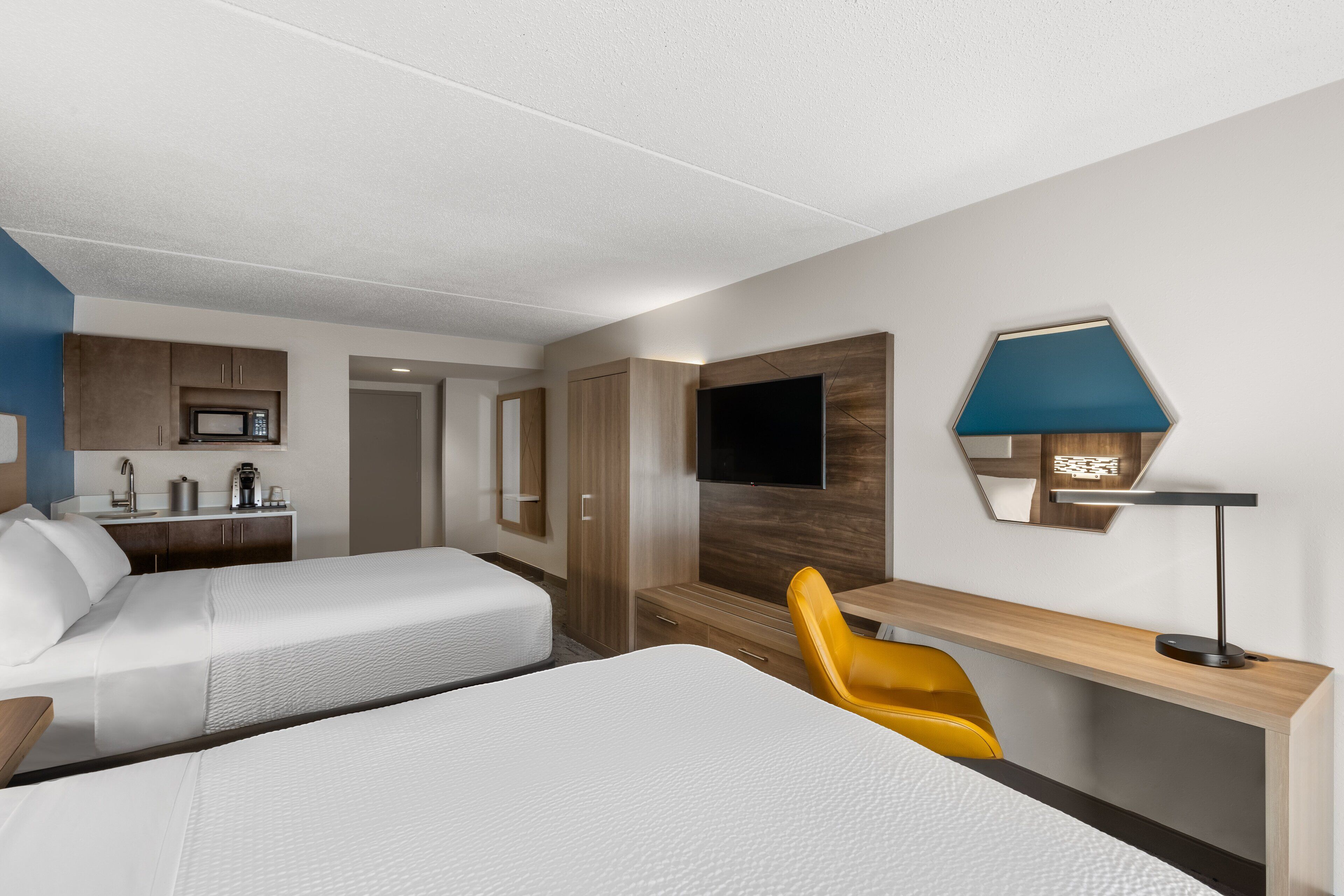 A hotel room with two beds, a desk, a chair, a TV, and a kitchenette.