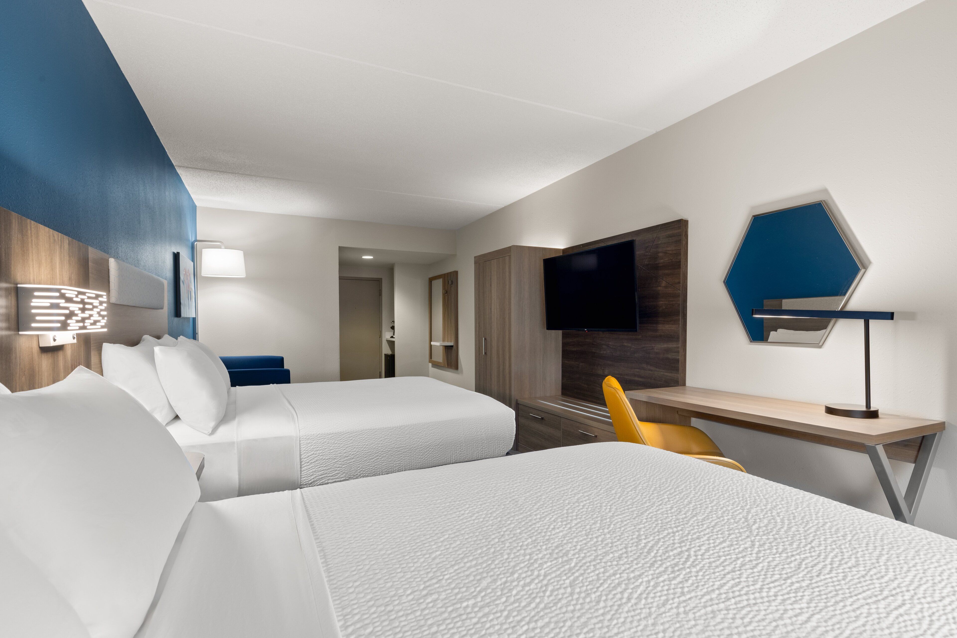 Quarto de hotel com duas camas, uma escrivaninha, uma cadeira, televisão e uma luminária na parede.
