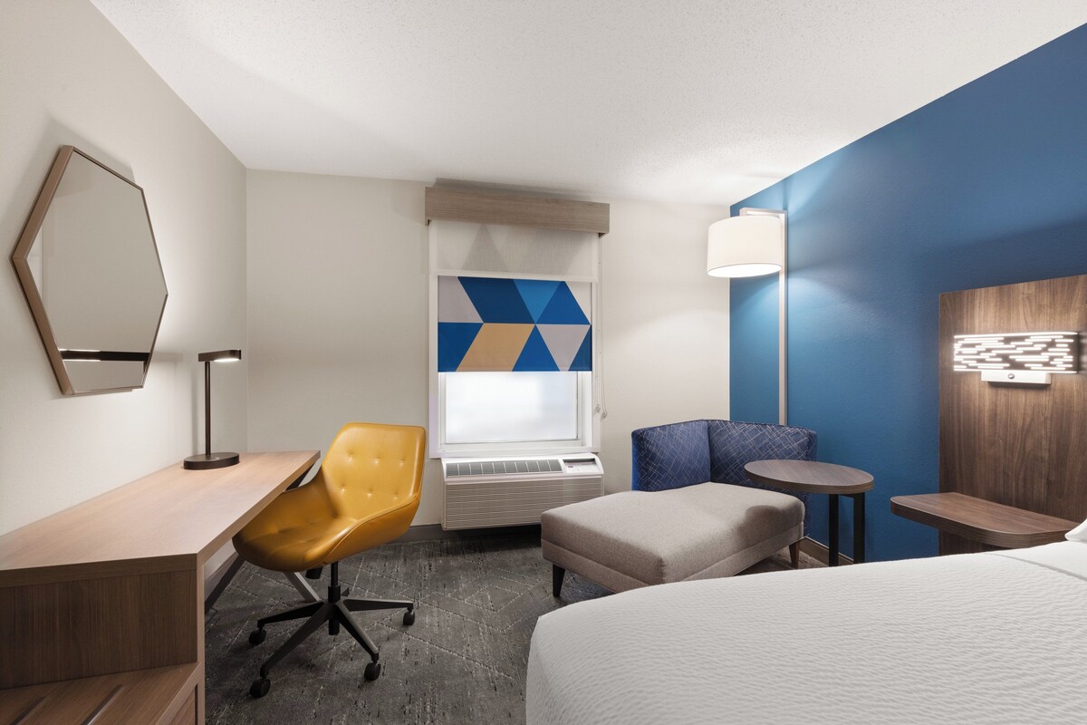 A hotel room with a bed, a desk, a chair, a small table, a lamp, and a window with a geometric view.