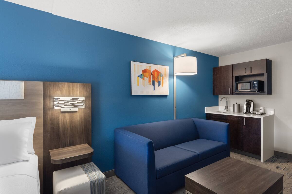 Ein modernes Hotelzimmer mit blauer Akzentwand, einer Couch, einem Bett, einem Nachttisch, einer Lampe und einer Küchenzeile.