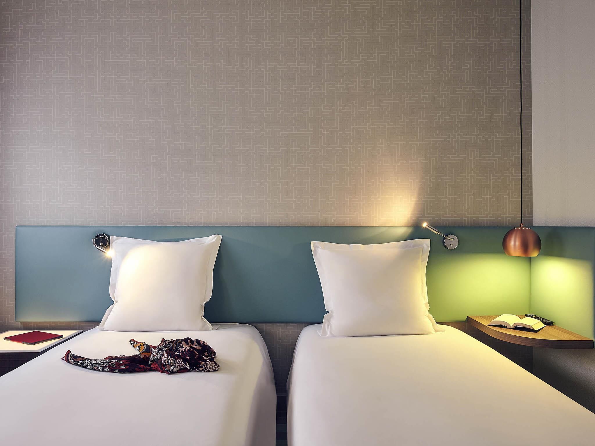Ein Hotelzimmer mit zwei Betten, einem Nachttisch mit Lampe und einem kleinen Tisch mit Buch und Fernbedienung.