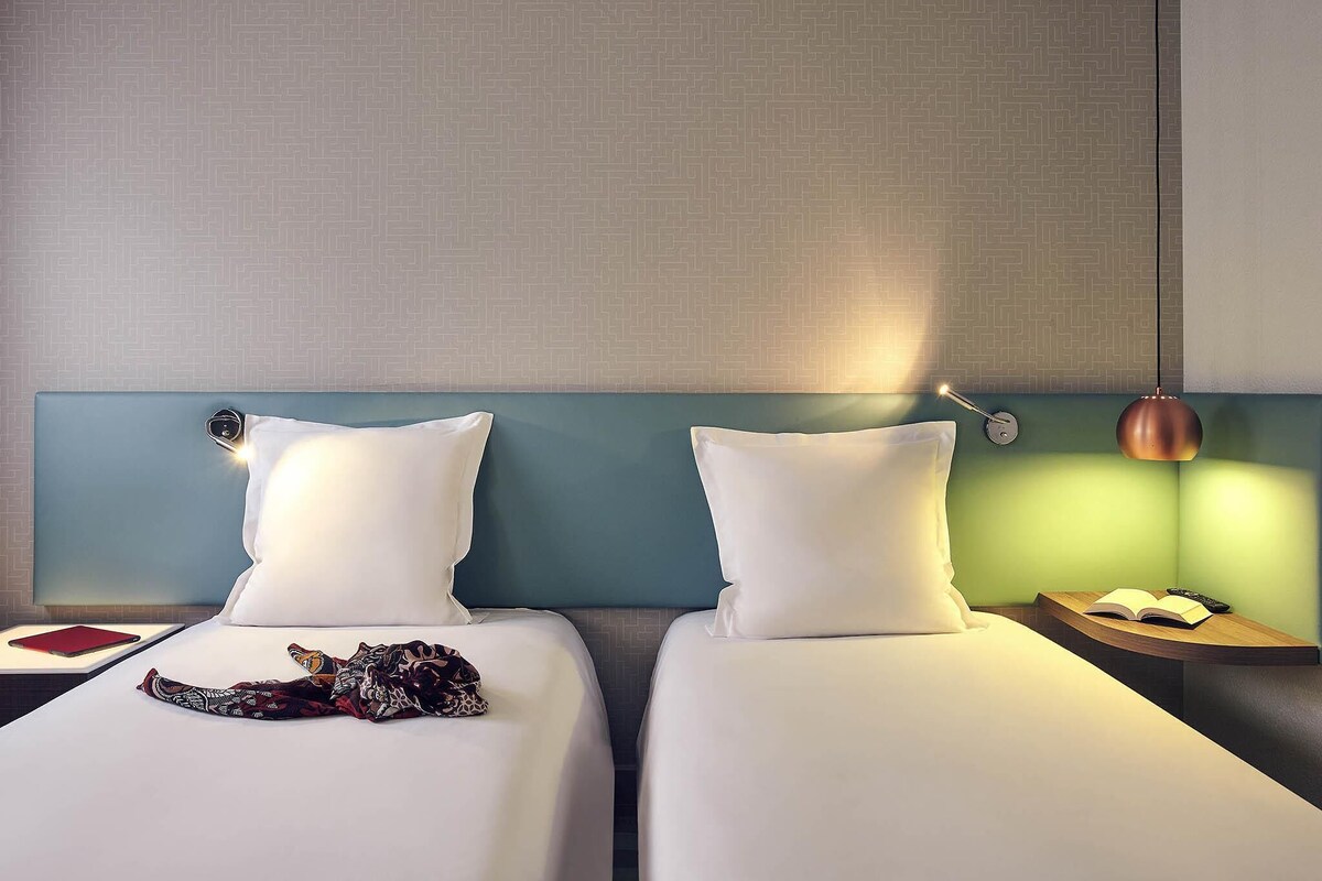 A hotel room with two beds, a nightstand with a lamp, and a small table with a book and remote control.