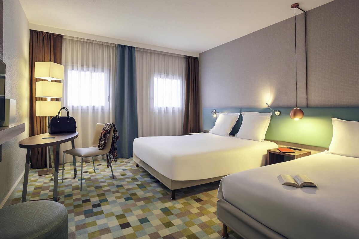 A hotel room with two beds, a desk, a chair, and a lamp.