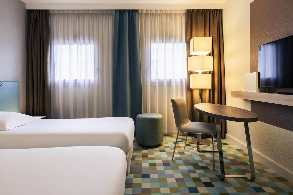 A hotel room with two beds, a desk, a chair, a TV, and a lamp.